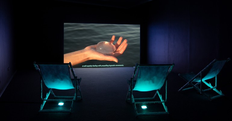 Kristina Õllek & Kert Viiart-Õllek, As the Earliest Carrier Emerges, 2024. Video installation, 4K video 10’ 10 min, beach chairs, digital prints on textile, clay, silicone, blue filter, LED lights. Photo by Kristina Õllek