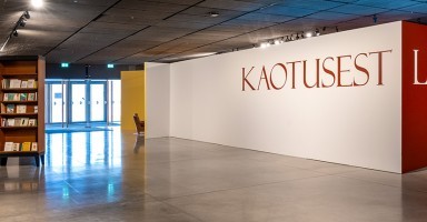 Kolmnurga moodustavatel seintel saab lugeda näituse pealkirja valgel ja punasel taustal. Foto vasakus nurgas on näha eesti pagulaskirjanike teostega täidetud raamaturiiulit. Taamal paistab läbi klaasuste taevas.